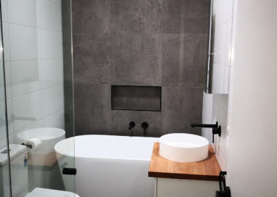 bathroom, with shower, toilet, bath against the wall, inbuilt shelf in wall with black accents, light wooden vanity, and mirror cabinet on the wall